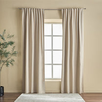 Beige curtains hanging on a window with a neutral wall and plant in the corner.