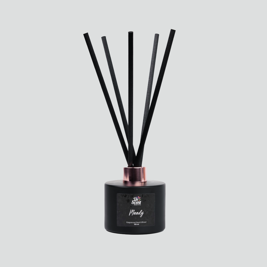 A black reed diffuser with a round base and several vertical reeds, with a copper-colored lid and a label that reads 'Woodly'.