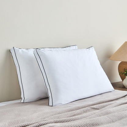 Two white pillows with black piping on a bed against a beige wall.