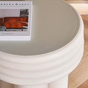 Round white table with a gray cushion and a magazine on a wooden floor.