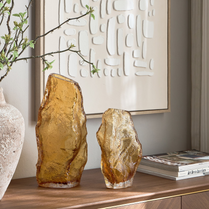 Two amber-colored stone sculptures on a wooden surface with a decorative wall art piece in the background.
