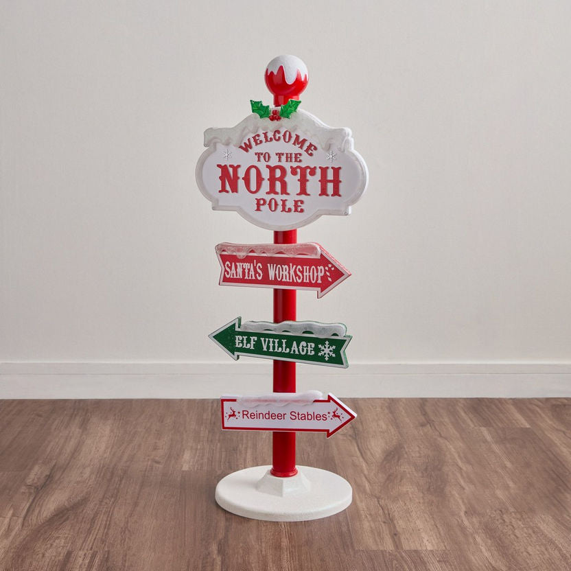 Christmas-themed decorative sign with directions to the North Pole, Santa's Workshop, Elf Village, and Reindeer Stables on a wooden floor.