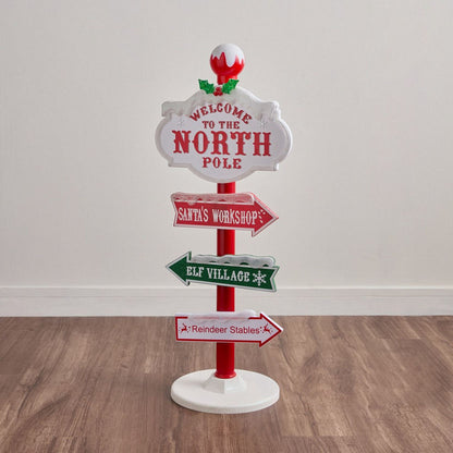 Christmas-themed decorative sign with directions to the North Pole, Santa's Workshop, Elf Village, and Reindeer Stables on a wooden floor.