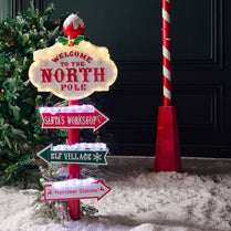 Christmas-themed signpost with directions to the North Pole, Santa's Workshop, Elf Village, and Reindeer Stables in front of a black door.