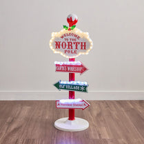 Decorative Christmas sign with directions to the North Pole, Santa's Workshop, Elf Village, and Reindeer Stables on a wooden floor.
