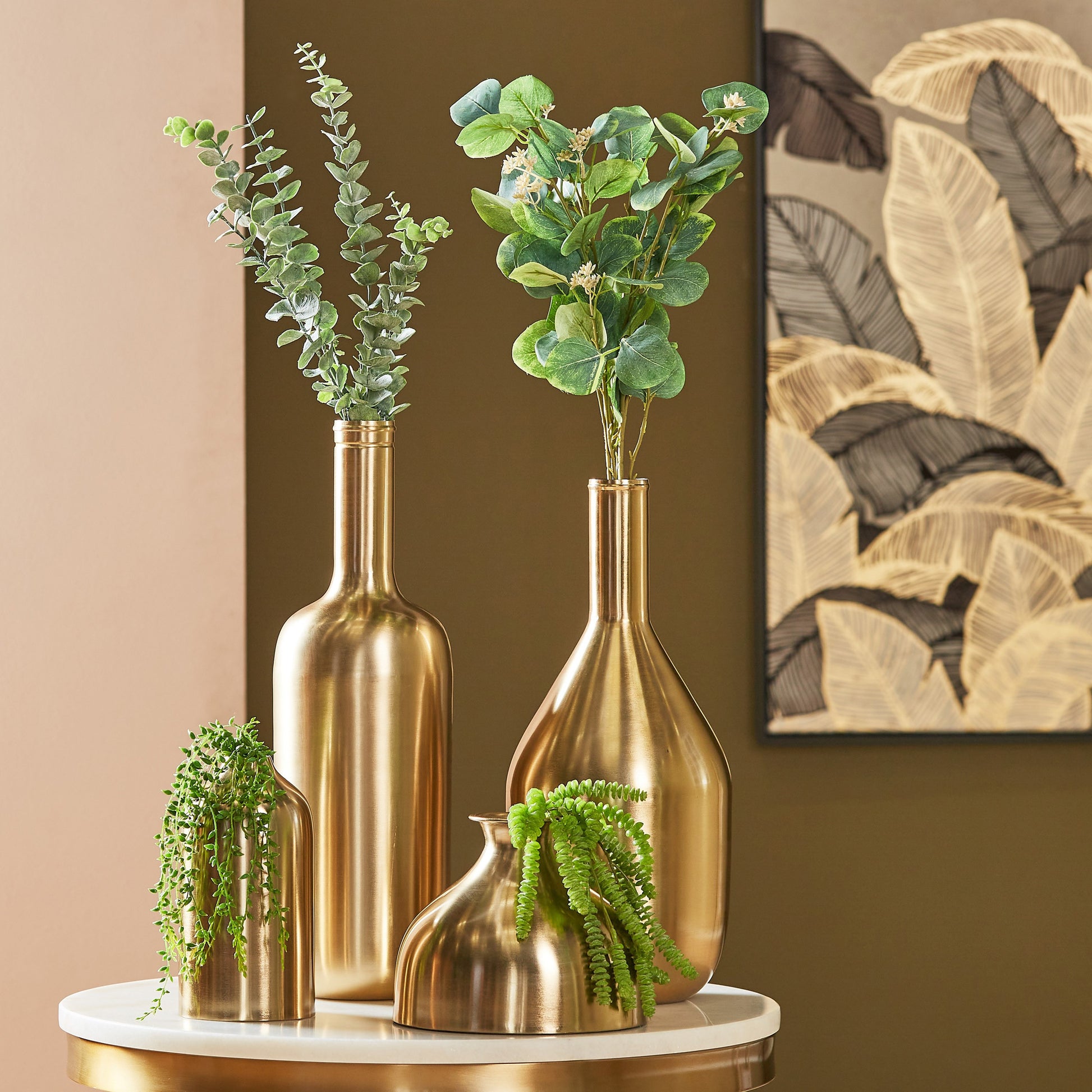 Gold vases with green plants on a marble surface against a brown wall with abstract art.