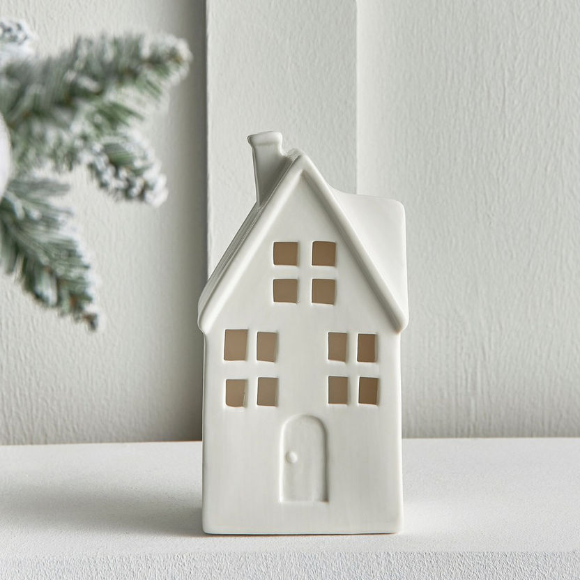 White ceramic house-shaped lantern on a white surface with a light gray wall and greenery in the background.