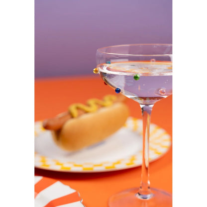 Hot dog with mustard on a plate next to a glass of water with colorful candies on an orange surface.