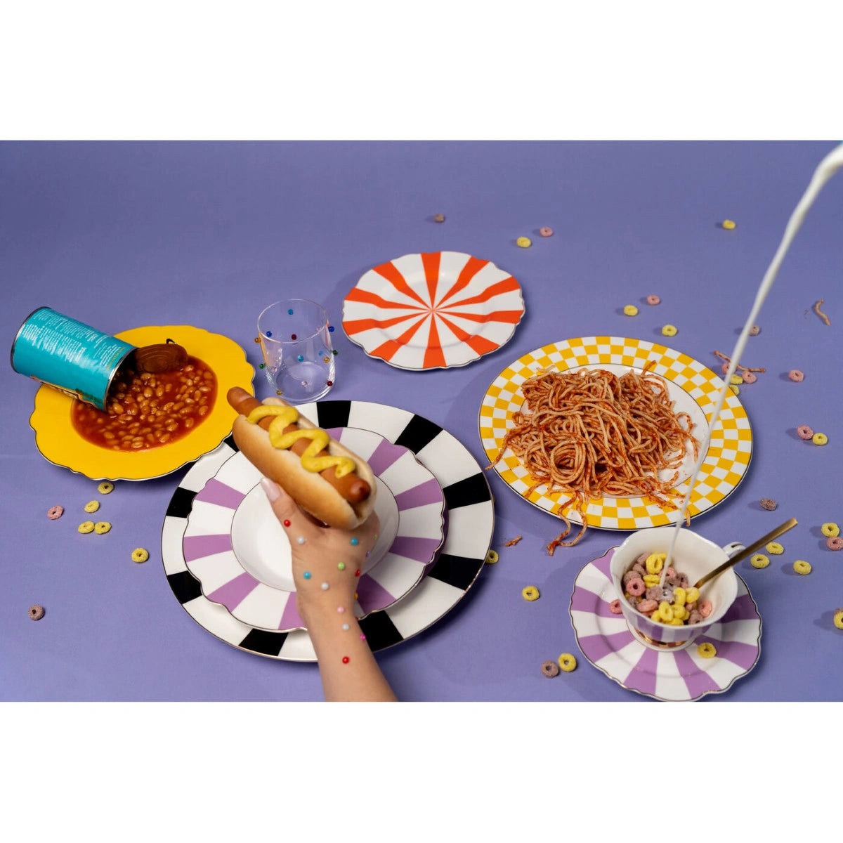 Colorful table setting with plates of food and a hand holding a hot dog on a purple background.