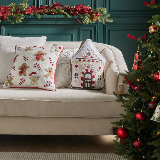 Decorative pillows with gingerbread men and a house design on a beige sofa next to a Christmas tree.