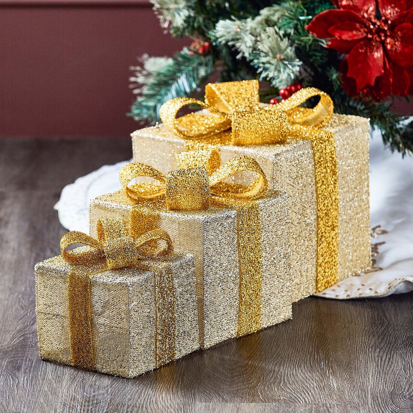 Stack of gold and silver glittery presents with gold ribbons on a wooden surface, Christmas tree in the background.