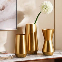 Three gold vases on a marble surface with a white flower and books in the background.