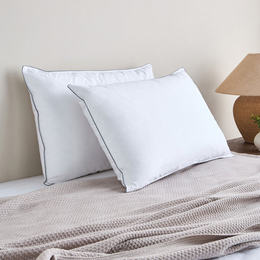 Two white pillows on a bed with a beige blanket and lamp in the background.