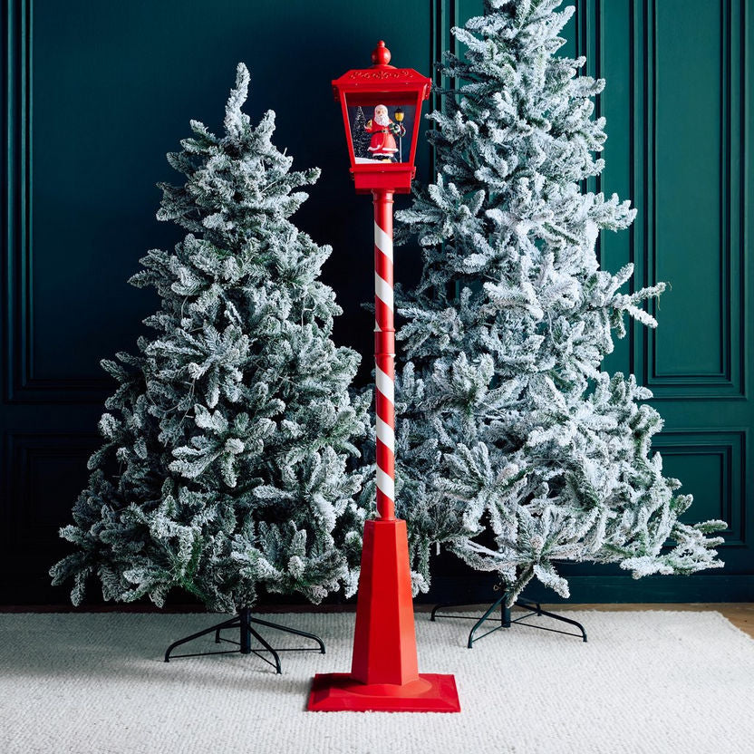 Red decorative lamp post with a Santa figure between two Christmas trees against a dark green wall.