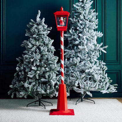 Red decorative lamp post with a Santa figure between two Christmas trees against a dark green wall.