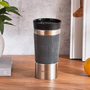 Gray travel mug with black lid on a wooden surface with a plant and books in the background