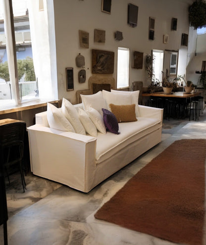 Living room with a white sofa, decorative pillows, and a dining area in the background. New York Collection Sofa – Live in the Now | Vella Concept