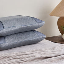Two blue textured pillows stacked on a textured surface with a lamp in the background.