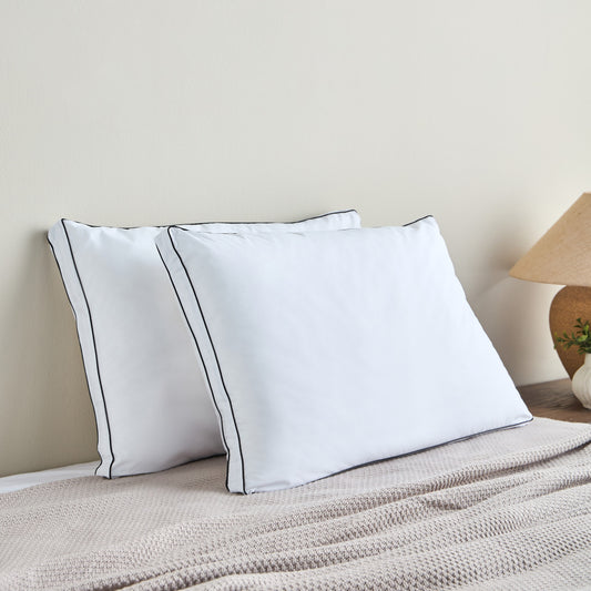 Two white pillows with black piping on a bed against a beige wall.
