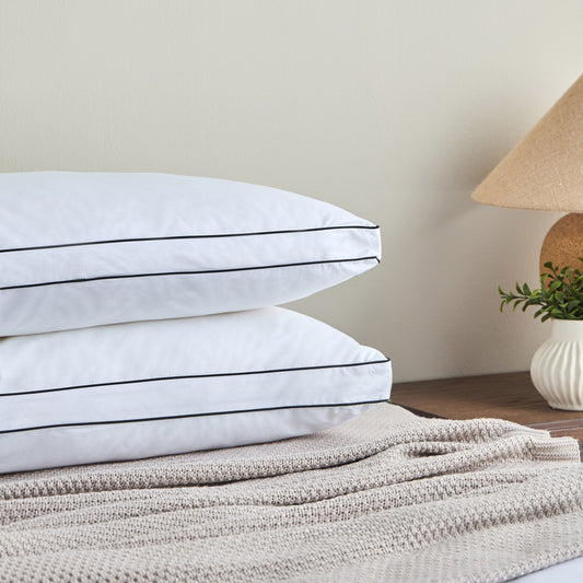Stack of white pillows with black stripes on a bed with beige bedding and a lamp in the background.