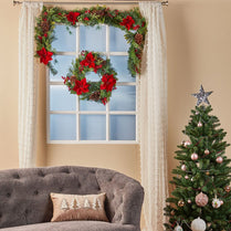 Decorative wreath with red flowers and greenery above a window, with a Christmas tree on the right.
