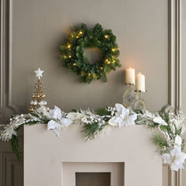 Decorative Christmas mantel with wreath, garland, and candles on a neutral wall.