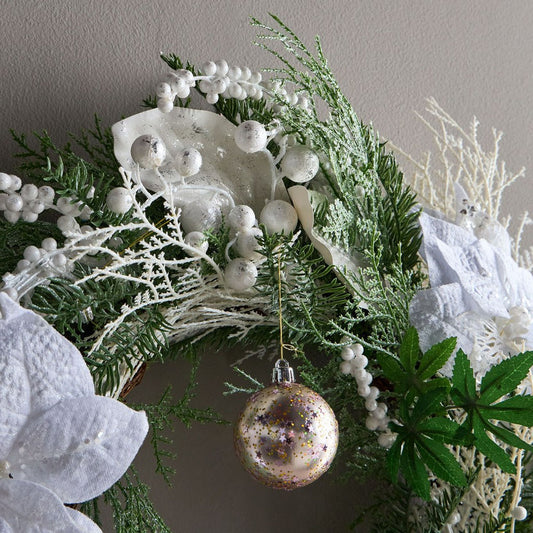 Decorative wreath with greenery, white flowers, and a gold ornament on a neutral background