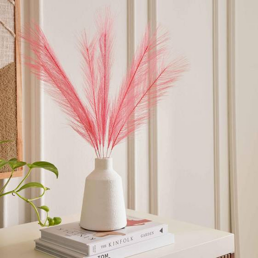 White vase with pink dried grasses on a stack of books against a neutral wall.
