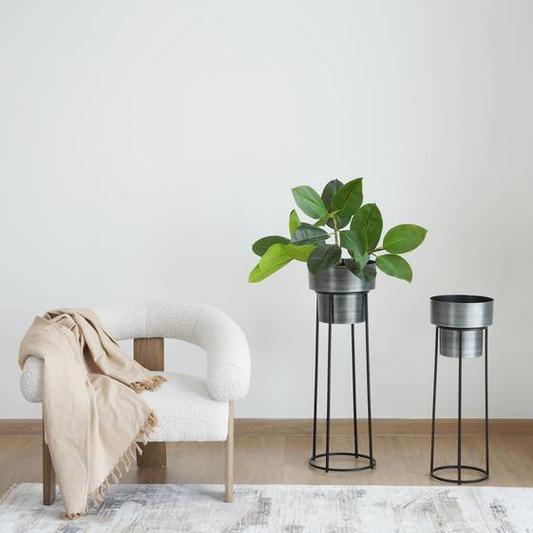 White armchair with a beige blanket and two metal plant stands with green plants on a light wooden floor.