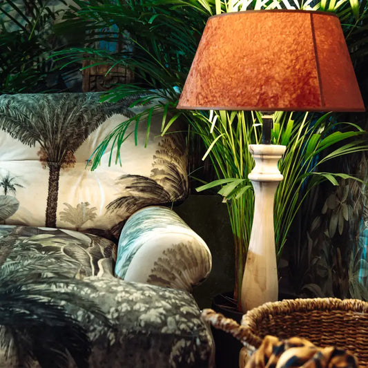 A cozy living room corner featuring the Couch Lucania Cythera, a white table lamp with a brown shade, lush green plants in the background, and handmade beech wood furniture woven seamlessly into the inviting setting.