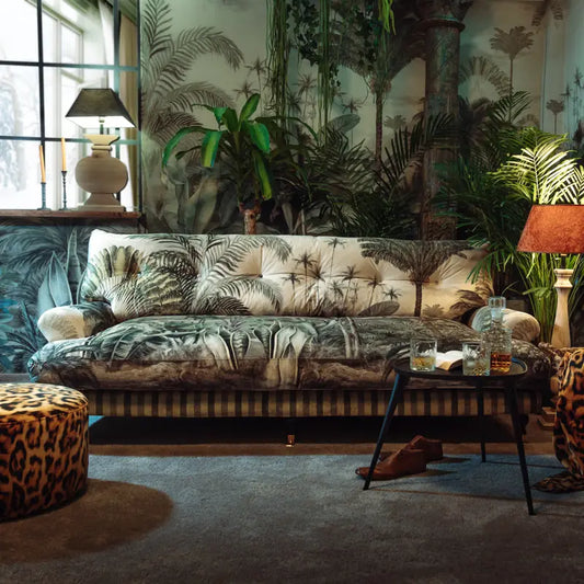 A cozy living room with the Couch Lucania Cythera, leafy plants, a patterned ottoman, and handmade beech wood furniture. Warm table lamp lighting and tropical wallpaper create a lush, inviting atmosphere.